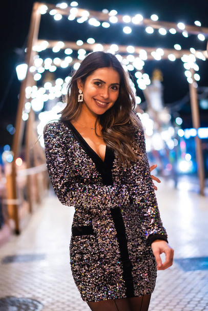 Christmas lifestyle. Portrait a young brunette Caucasian woman in a fashionable dress with sequins, smiling in the Christmas lights of the city, vertical photo - 写真・画像