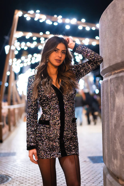 Christmas lifestyle. Brunette Caucasian model wearing fashionable dress with sequins, portrait in city christmas lights, vertical photo - Foto, afbeelding