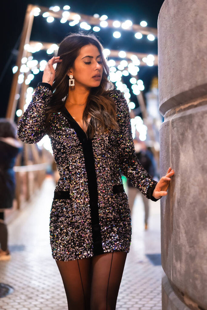 Christmas lifestyle. Brunette Caucasian model wearing a fashionable sequined dress, portrait in city christmas lights - Фото, зображення