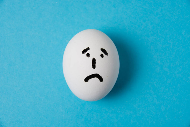 Ένα αυγό με λυπημένο πρόσωπο. Απομόνωση σε μπλε φόντο. - Φωτογραφία, εικόνα