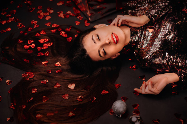 Top view of a girl in a shiny dress on the floor in confetti in the form of hearts and gifts. - Foto, imagen