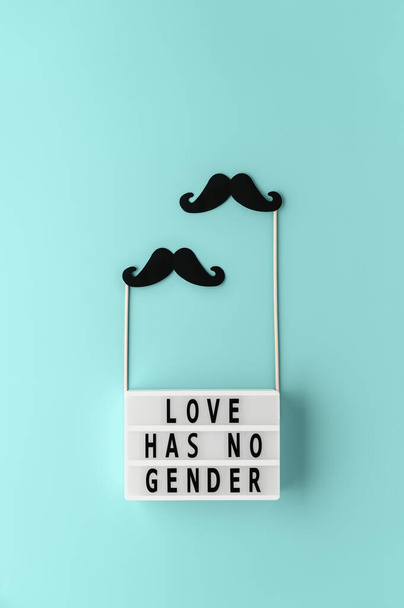 Lightbox with text LOVE HAS NO GENDER, black paper party accessories mustache on pastel cyan background. Concept Homosexuality, gay love. Top view, creative flat lay. Vertical orientation - Photo, Image