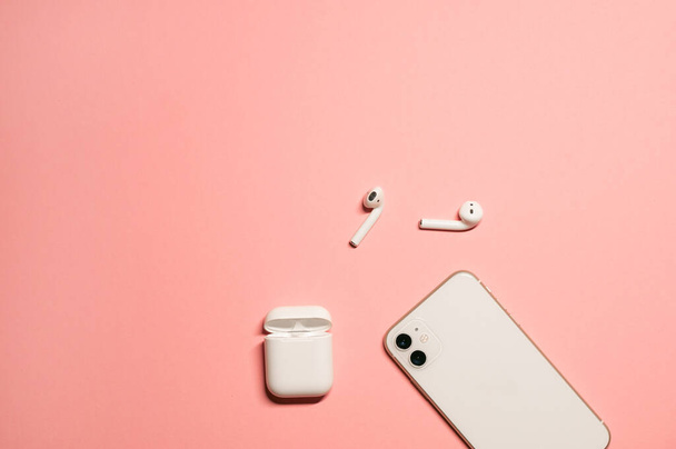 Simple flat lay with a stylish white smartphone and modern wireless earphones with a charging case on a pastel pink background. - Photo, Image