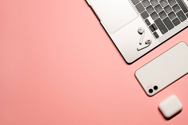 Красочный плоский снимок серебристо-серого ноутбука, белого смартфона, беспроводных наушников с зарядным чехлом на розовом фоне. - Фото, изображение