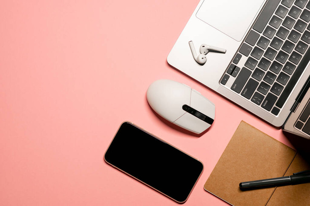 Diseño horizontal sobre un fondo rosa pastel con un ordenador portátil moderno, ratón inalámbrico, teléfono inteligente con pantalla táctil, auriculares, portátil y un bolígrafo. - Foto, Imagen
