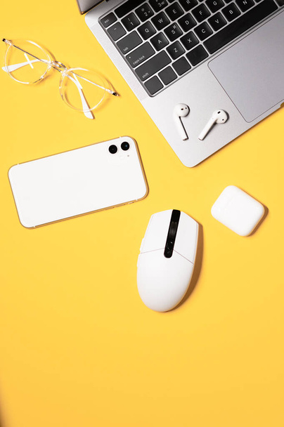 Κάθετη διάταξη με ένα λευκό ποντίκι υπολογιστή, σύγχρονο γκρι φορητό υπολογιστή με μαύρο πληκτρολόγιο, λευκό smartphone, γυαλιά, και ασύρματα ακουστικά σε ένα πολύχρωμο κίτρινο φόντο. - Φωτογραφία, εικόνα