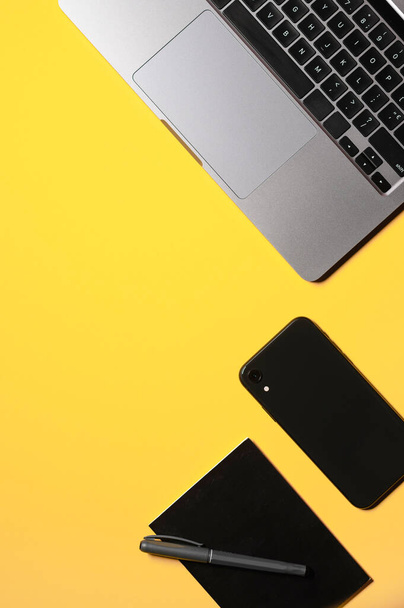 Gri dizüstü bilgisayarı, siyah akıllı telefonu, not defteri ve izole edilmiş sarı arka planda bir kalemi olan dikey düz bir kompozisyon.. - Fotoğraf, Görsel
