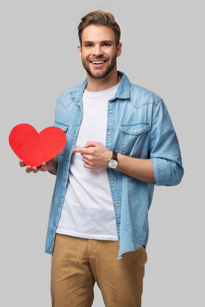 Portrét šarmantní muž držet ruční papír srdce ve tvaru karty nosit ležérní styl košile izolované přes šedé pozadí - Fotografie, Obrázek
