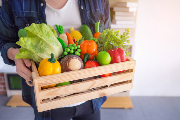 Lähikuva kuva käsissä nuori mies pitää kori laatikko puinen värikkäitä vihanneksia ja hedelmiä orgaaninen valmistautua elintarvikkeita keittiössä. Terveyden ja terveydenhuollon käsite. - Valokuva, kuva