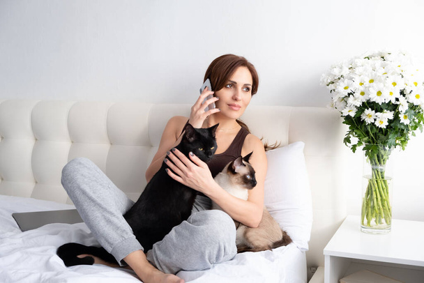 mujer joven hablando por teléfono con sus dos gatos en el dormitorio blanco con flores - Foto, Imagen