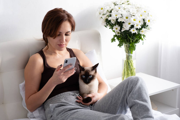 jonge vrouw met behulp van telefoon met haar Siamees kat op slaapkamer met bloemen - Foto, afbeelding