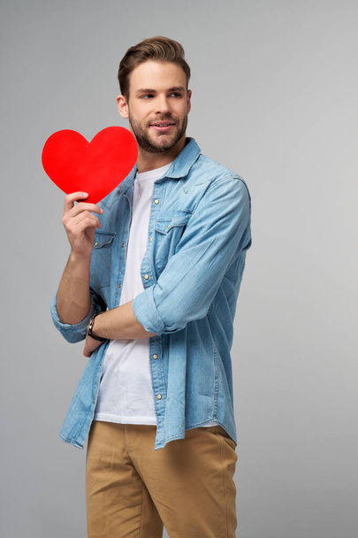 Portrét šarmantní muž držet ruční papír srdce ve tvaru karty nosit ležérní styl košile izolované přes šedé pozadí - Fotografie, Obrázek