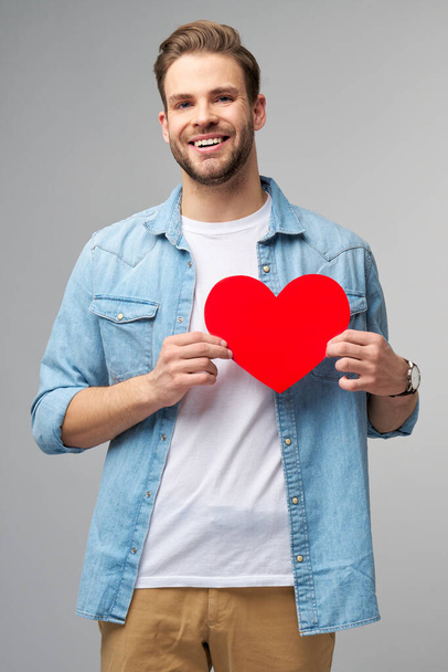 Portret van een charmante man houden hand papier hartvormige kaart dragen casual stijl shirt geïsoleerd over grijze achtergrond - Foto, afbeelding