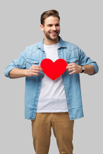 Retrato de hombre encantador de la mano de papel en forma de corazón de la tarjeta de desgaste camisa de estilo casual aislado sobre fondo gris - Foto, Imagen