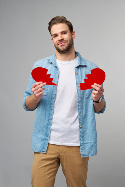 handsome young man holding broken paper red valentine heart standing over grey background - Фото, изображение