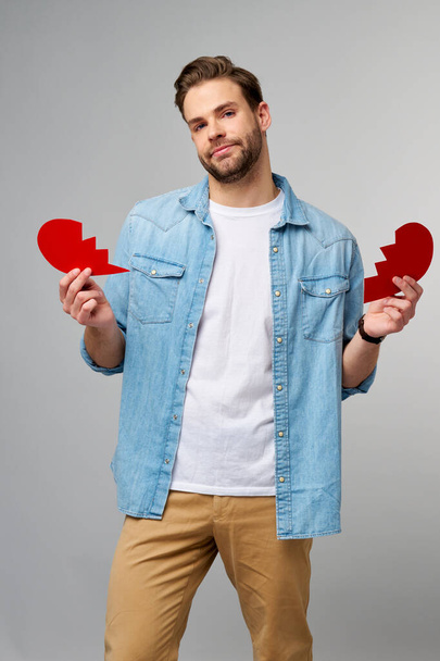 pohledný mladý muž drží zlomený papír červený valentine srdce stojící nad šedým pozadím - Fotografie, Obrázek