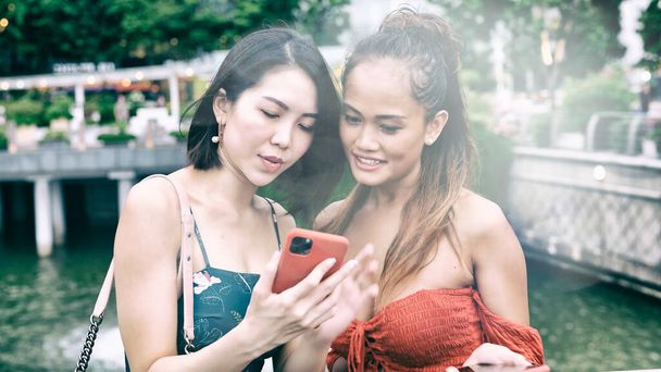 Дві красиві молоді азіатські жінки розмовляють, посміхаються і розслабляються, ходять по міських вулицях, дивлячись на свої смартфони
. - Фото, зображення