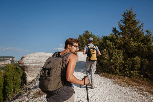 Dos hombres caminando en la ladera rocosa llevando mochilas usando palos de trekking. Valle de las montañas Vista al lado de la gente. viaje de verano a la naturaleza. Caminante en segundo plano. - Foto, imagen