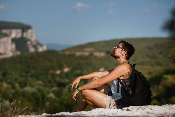 Giovane escursionista di sesso maschile con zaino rilassarsi sulla cima di una montagna durante il calmo tramonto estivo scenario da vacanza. Uomo viaggiatore stanco che riposa sulla collina. - Foto, immagini