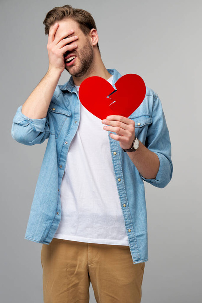 handsome young man holding broken paper red valentine heart standing over grey background - Fotografie, Obrázek