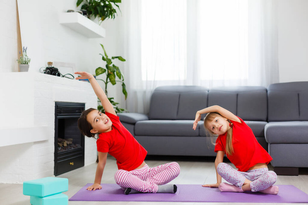 Дві маленькі дівчата практикують йогу, розтягуються, пристосовуються до відео на ноутбуці. Дистанційна онлайн-навчання, аеробіка вдома. Здоровий спосіб життя, коронавірус, залишайся вдома. Дитячий спортивний домашній карантин
. - Фото, зображення