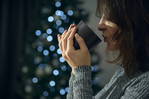 Egy fiatal nő megiszik egy csésze teát vagy forró csokit, hogy felmelegedjen a tél hidegétől. A háttérben a karácsonyfa fényei bokeh hatást keltenek, hogy megünnepeljék a szent ünnepséget.. - Fotó, kép