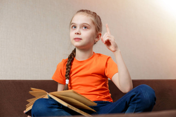 Cute Little Girl Reading a Book on Sofa in lotus Position at Home. Holds Forefinger up Ideas. - Photo, Image