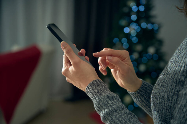 A young woman use the smartphone to socialize, wishes and celebrate with friends online from her living room. Smartphone mockup is on a bokeh effect background.  - Photo, Image