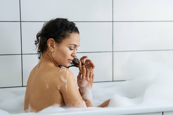 Sise θέα μιας όμορφης γυναίκας που χαλαρώνει στην μπανιέρα και πλένει σγουρά μαλλιά της - Φωτογραφία, εικόνα