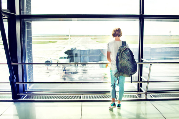 Ταξιδιωτικός τουρίστας στέκεται με αποσκευές βλέποντας στο παράθυρο του αεροδρομίου. Αγνώριστη γυναίκα με κοντά μαλλιά κοιτάζει το σαλόνι κοιτάζοντας αεροπλάνα στην πύλη επιβίβασης. Ταξιδιωτικός τρόπος ζωής. - Φωτογραφία, εικόνα