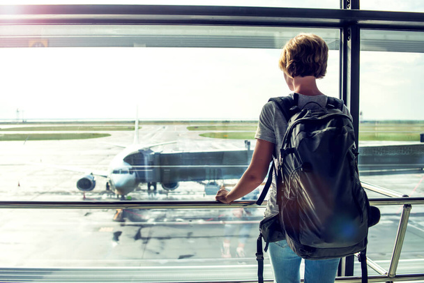 Ταξιδιωτικός τουρίστας στέκεται με αποσκευές βλέποντας στο παράθυρο του αεροδρομίου. Αγνώριστη γυναίκα με κοντά μαλλιά κοιτάζει σαλόνι κοιτάζοντας αεροπλάνα στην πύλη επιβίβασης πριν από την αναχώρηση. Ταξιδιωτικός τρόπος ζωής. - Φωτογραφία, εικόνα
