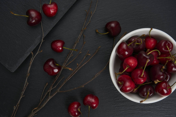 Un grupo de cerezas rojas y maduras en un tazón de cerámica blanca. Como fondo un plato de pizarra negra con las ramas secas del árbol. - Foto, Imagen