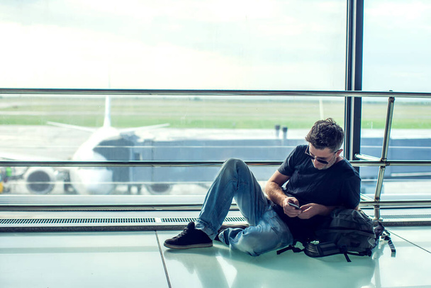 Νεαρός άνδρας ελέγχει το τηλέφωνό του ενώ περιμένει την πτήση του στο αεροδρόμιο. Ταξιδιωτική έννοια - Φωτογραφία, εικόνα