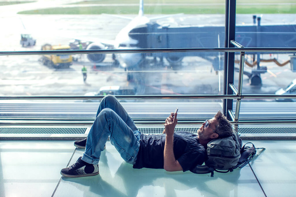 Nuori mies tsekkaa puhelintaan odottaessaan lentoa lentokentällä. Matkustuskäsite - Valokuva, kuva