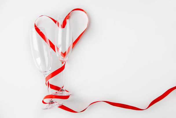 Two empty wine glasses with a red ribbon laid out in the shape of a heart on a white background. Flat lay with copyspace - Foto, afbeelding