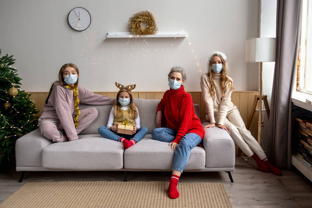 Coronavirus-Pandemie Weihnachten und Neujahr 2021 Konzept. Weibliche Familie trägt Schutzmasken und sitzt auf einer Couch und hält soziale Distanz im weihnachtlichen Wohnzimmer. - Foto, Bild