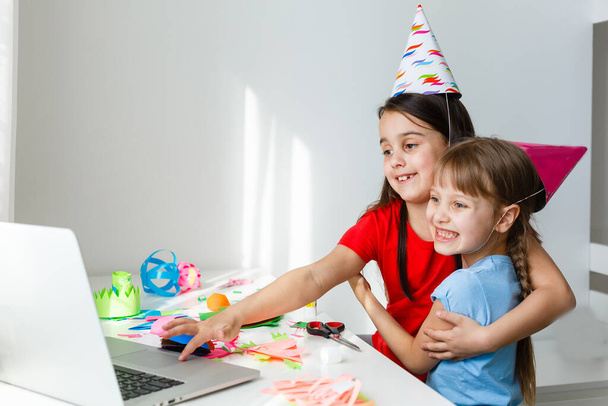 Детский онлайн день рождения. Маленькие девочки в платьях, шляпа празднуют праздник с друзьями. Конференция, видеозвонок на ноутбук, компьютер. Карантин, коронавирусная пандемия ковид-19. - Фото, изображение