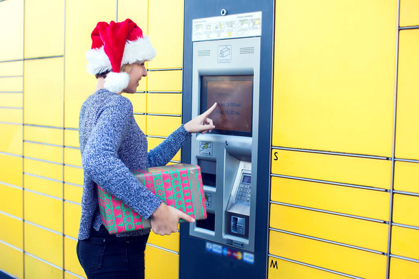 Nainen joulupukki hattu asiakas käyttää automaattista itsepalvelua post terminaali kone tai kaappi tallettaa paketin varastointiin - Valokuva, kuva