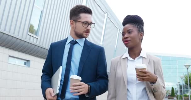 Mixed-races young businessman and businesswoman walking outdoor, holding coffee-to-go and talking. Multi ethnic businessman and businesswoman strolling the street and discussing business with drinks. - Footage, Video