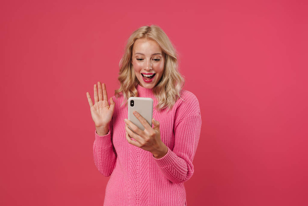 Όμορφη νεαρή ξανθιά κοπέλα με πουλόβερ που ποζάρει απομονωμένη σε ροζ φόντο, σε βιντεοκλήση μέσω κινητού τηλεφώνου - Φωτογραφία, εικόνα