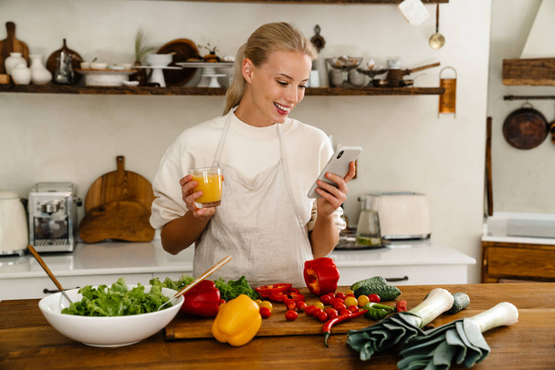 Beautiful happy woman using cellphone and drinking juice while making lunch in cozy kitchen - Valokuva, kuva