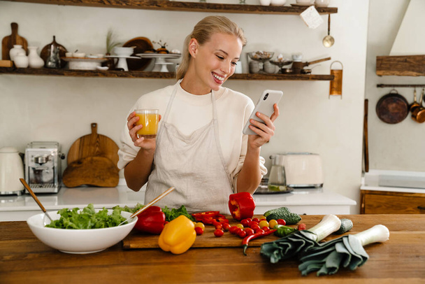 Beautiful happy woman using cellphone and drinking juice while making lunch in cozy kitchen - Фото, зображення