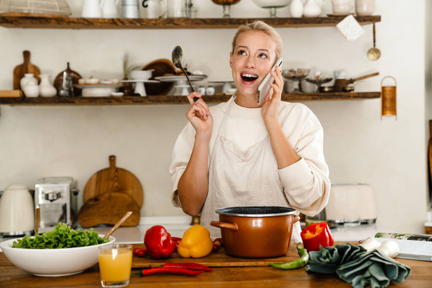 Beautiful surprised woman talking on cellphone while making lunch in cozy kitchen - Foto, afbeelding