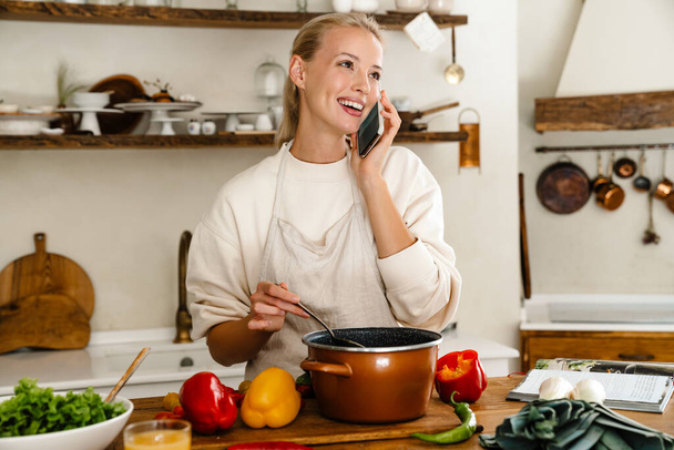 Beautiful happy woman smiling and talking on cellphone while making lunch in cozy kitchen - Фото, зображення
