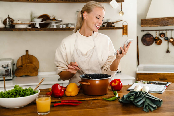Beautiful cheerful woman smiling and using cellphone while making lunch in cozy kitchen - Photo, Image