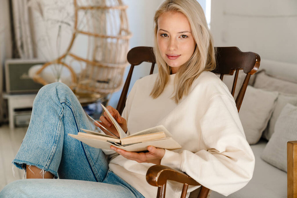 Krásná soustředěná žena čtení knihy, zatímco sedí v křesle v útulném pokoji - Fotografie, Obrázek