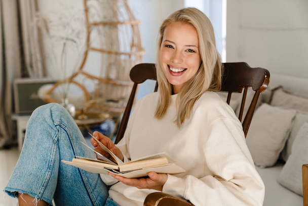 Beautiful happy woman smiling and reading book while sitting in armchair at cozy room - Foto, imagen