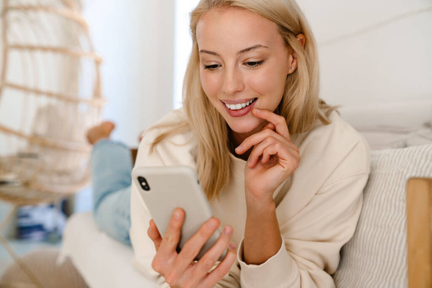Beautiful cheerful woman smiling and using cellphone while lying on couch in cozy room - Fotó, kép
