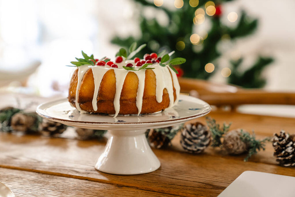 Großaufnahme eines Weihnachtskuchens mit Zuckerguss und Beeren auf dem Tisch drinnen, Weihnachtsbaum auf dem Hintergrund - Foto, Bild