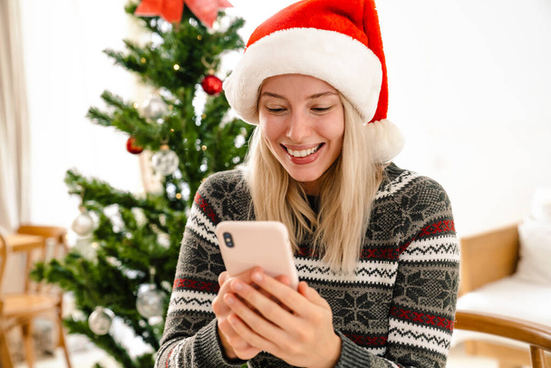 Photo of a young cheery woman in christmas santa hat indoors at home using mobile phone - Φωτογραφία, εικόνα
